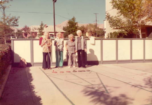 Four people standing on a sunlit driveway beside a white fence and house gate.