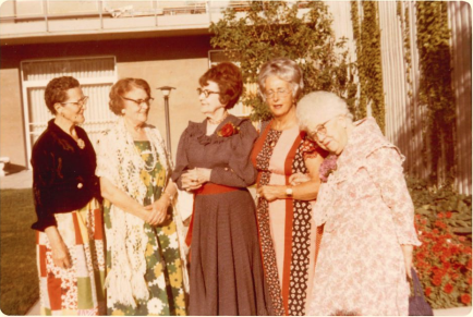 Five women standing together outdoors near a building and flowers, chatting in the sun.