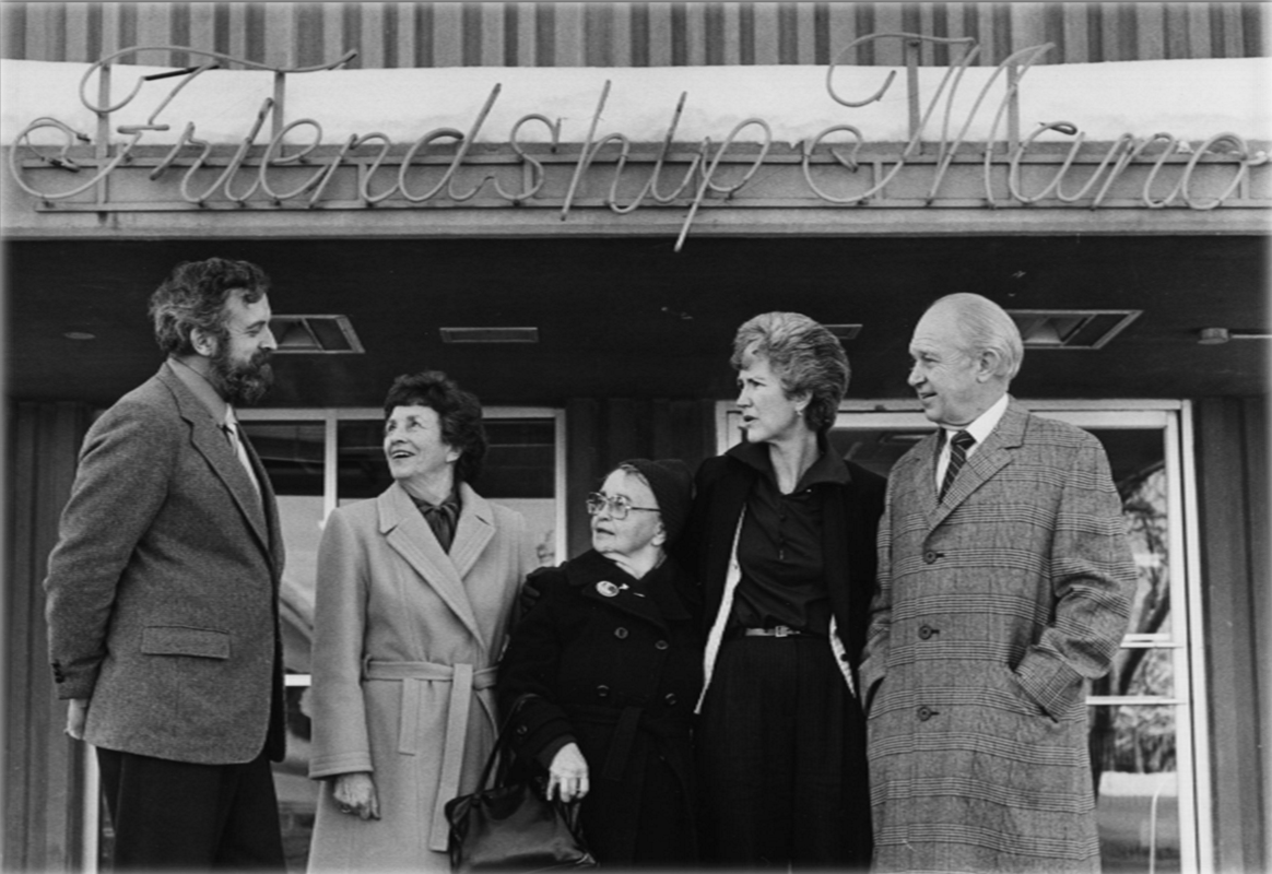 Five people stand outside a building under a sign, posing and smiling in black-and-white.