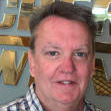 A man in a plaid shirt is smiling in front of a sign.