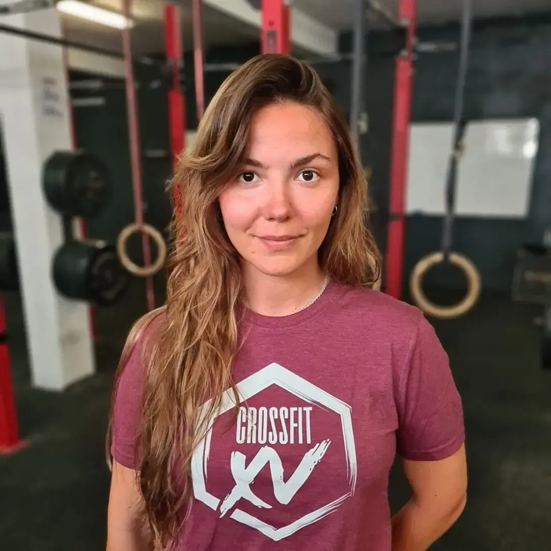 Woman in a maroon gym T-shirt standing in a fitness gym with rings and weights in the background