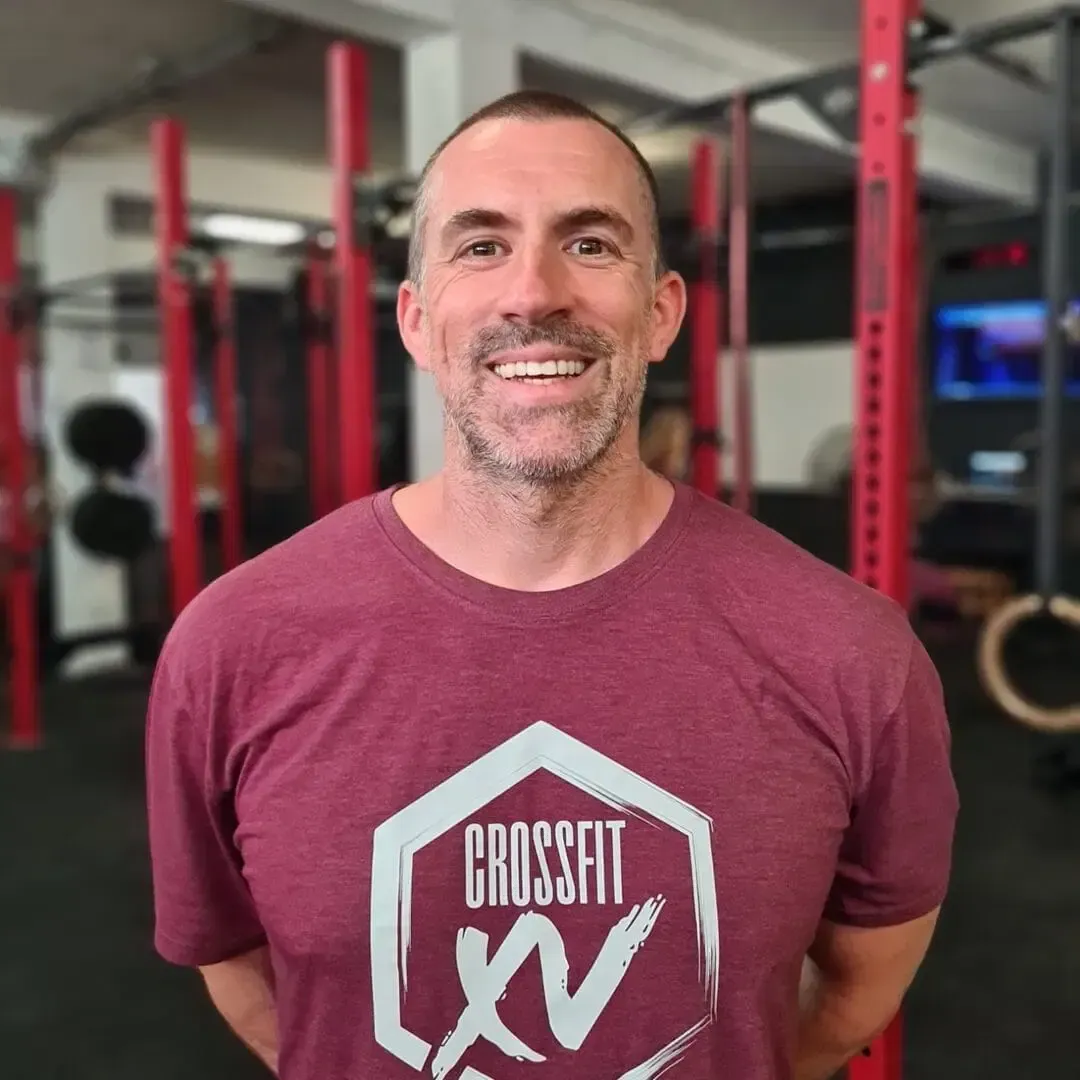 Smiling man in maroon CrossFit shirt standing in a gym with red racks behind him