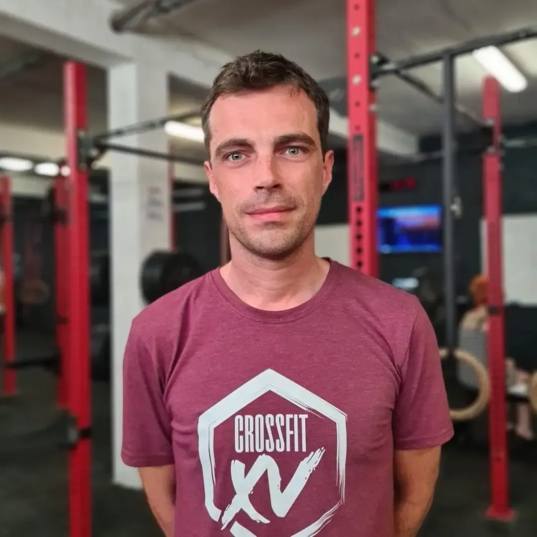 Person standing in a gym, wearing a maroon CrossFit shirt, with red equipment in the background.