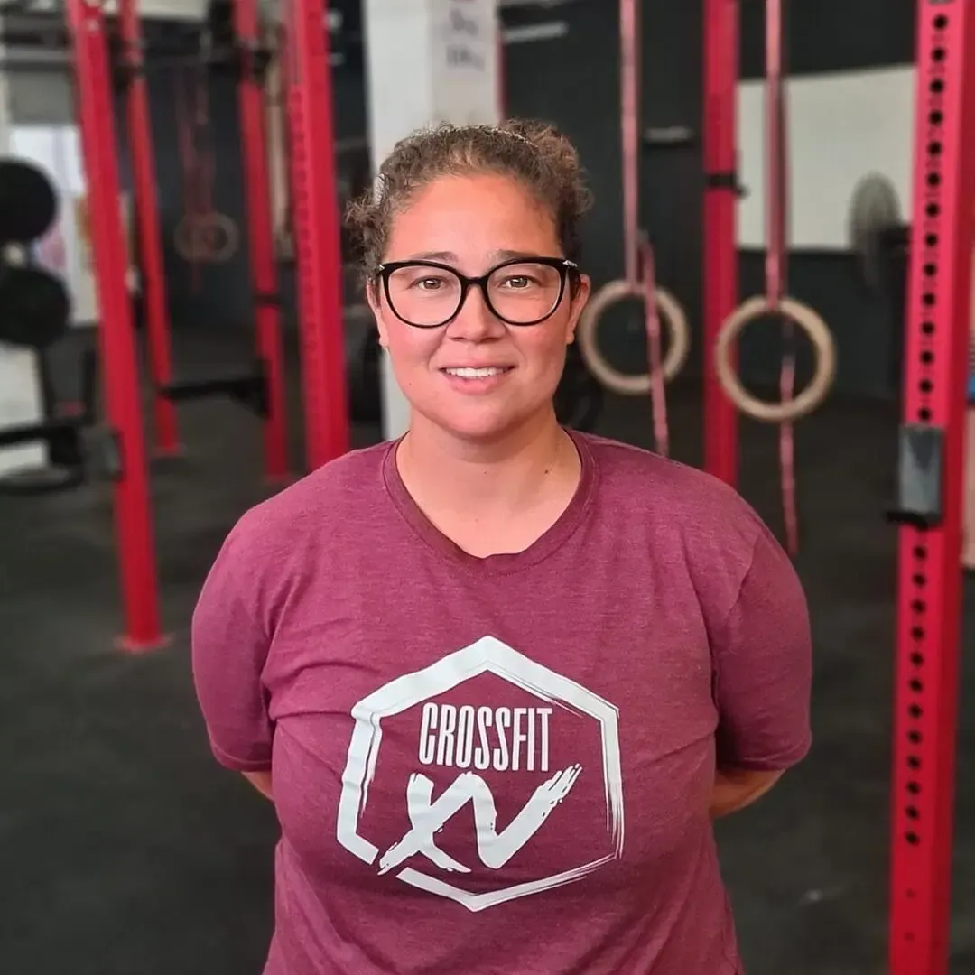 Woman in glasses and maroon CrossFit shirt standing in a gym with rings and red equipment behind her