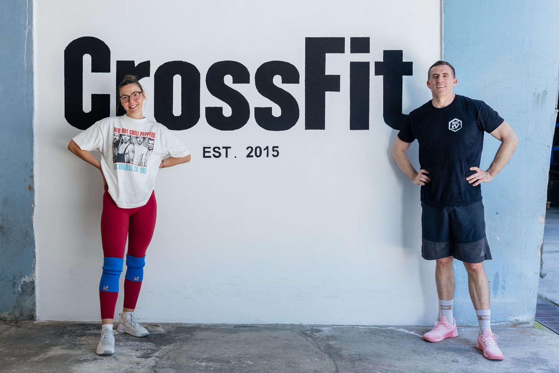 Two people standing in front of a CrossFit sign, hands on hips, in a gym setting.