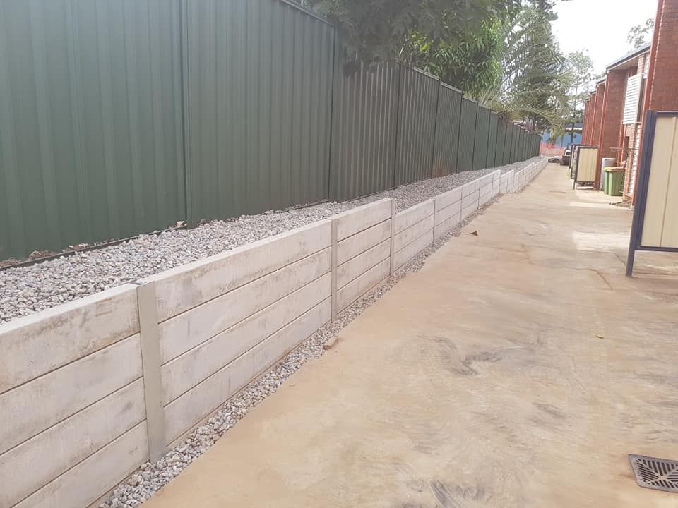 Concrete retaining wall with green metal fence and gravel along a paved alleyway.