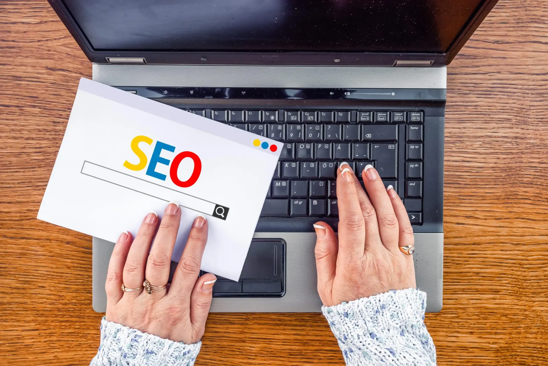 Person typing on a laptop keyboard, with a paper displaying “SEO” in a search bar.