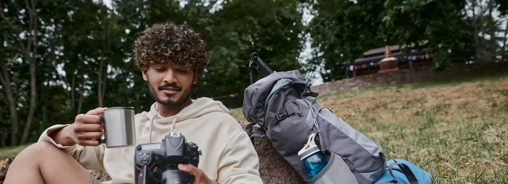A man is sitting in the grass with a backpack and a cup of coffee.