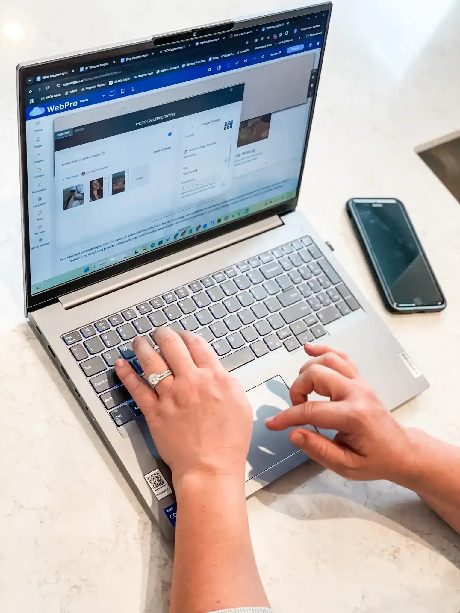 A person is typing on a laptop computer next to a cell phone.