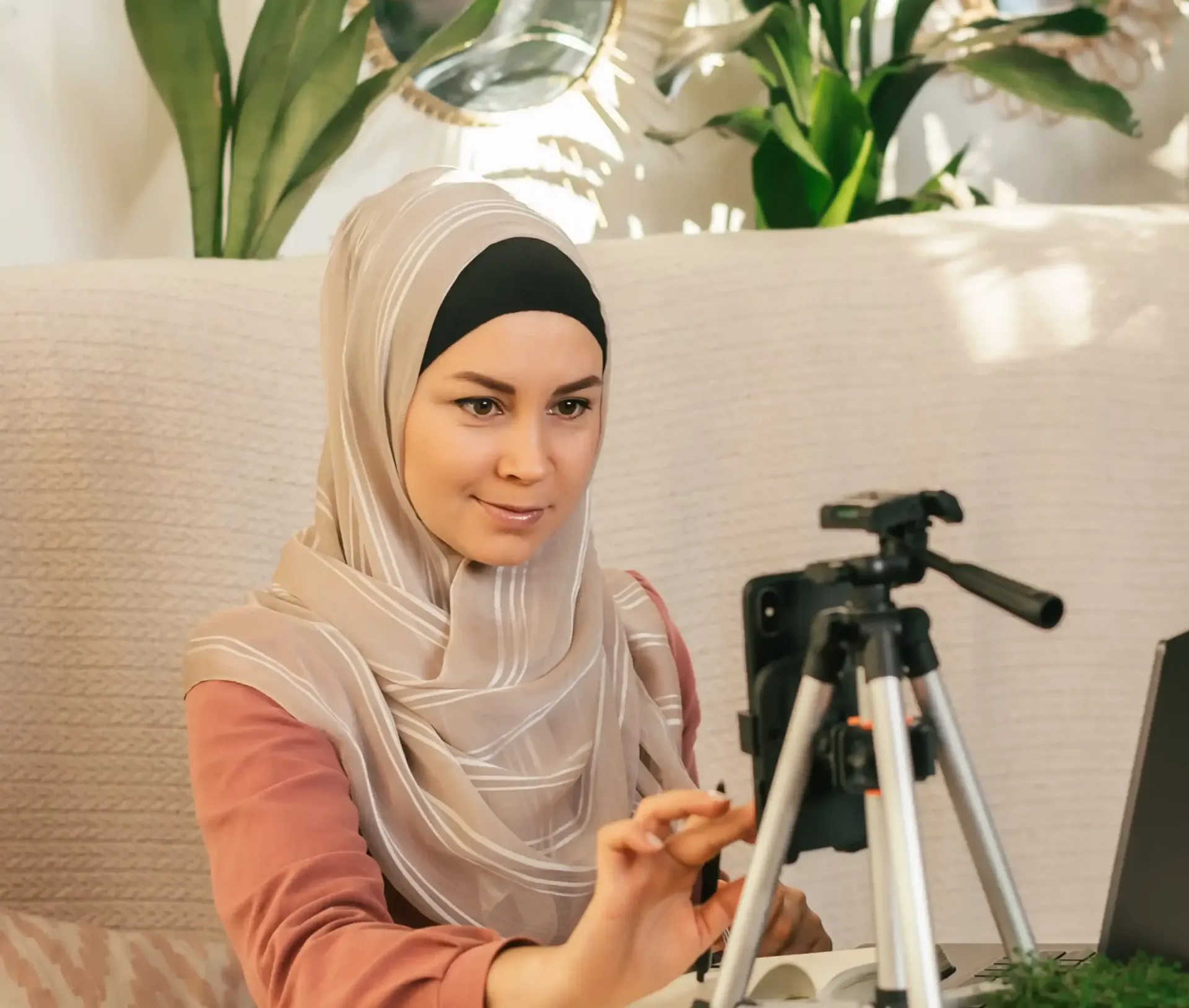A woman in a hijab is sitting in front of a laptop and a camera on a tripod creating content for her coaching business and SEO website.