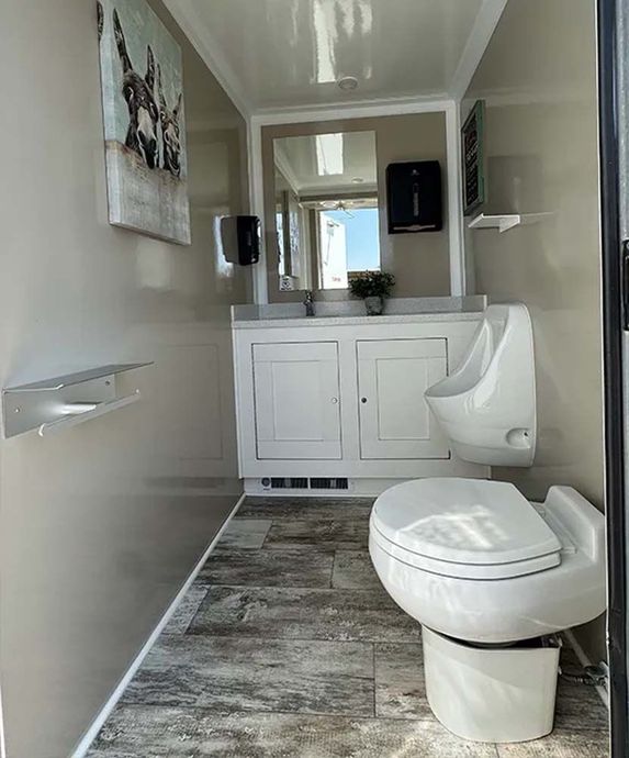 Interior of a luxury portable restroom with toilet, urinal, sink, and artwork.