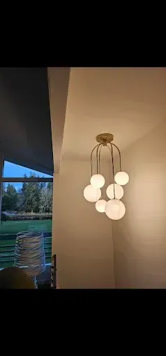 A modern chandelier with multiple glowing white glass orbs hangs from a metallic fixture in a room near a large window.