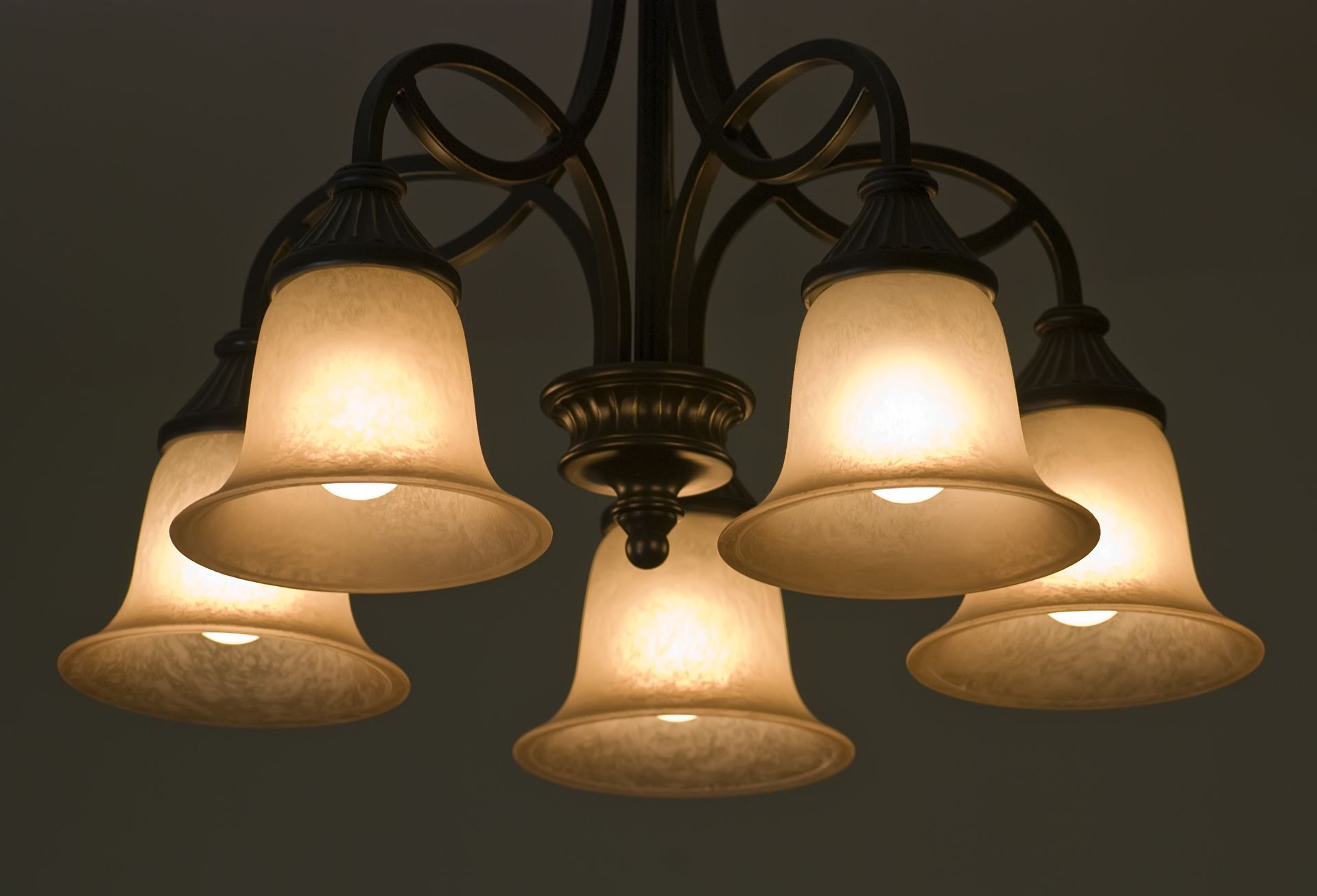 A five-light chandelier with frosted, bell-shaped glass shades and a dark bronze finish, illuminated against a dark background.