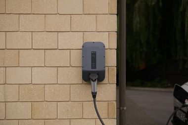 A grey electric vehicle charging station mounted on a light-colored brick wall with a charging cable plugged in.