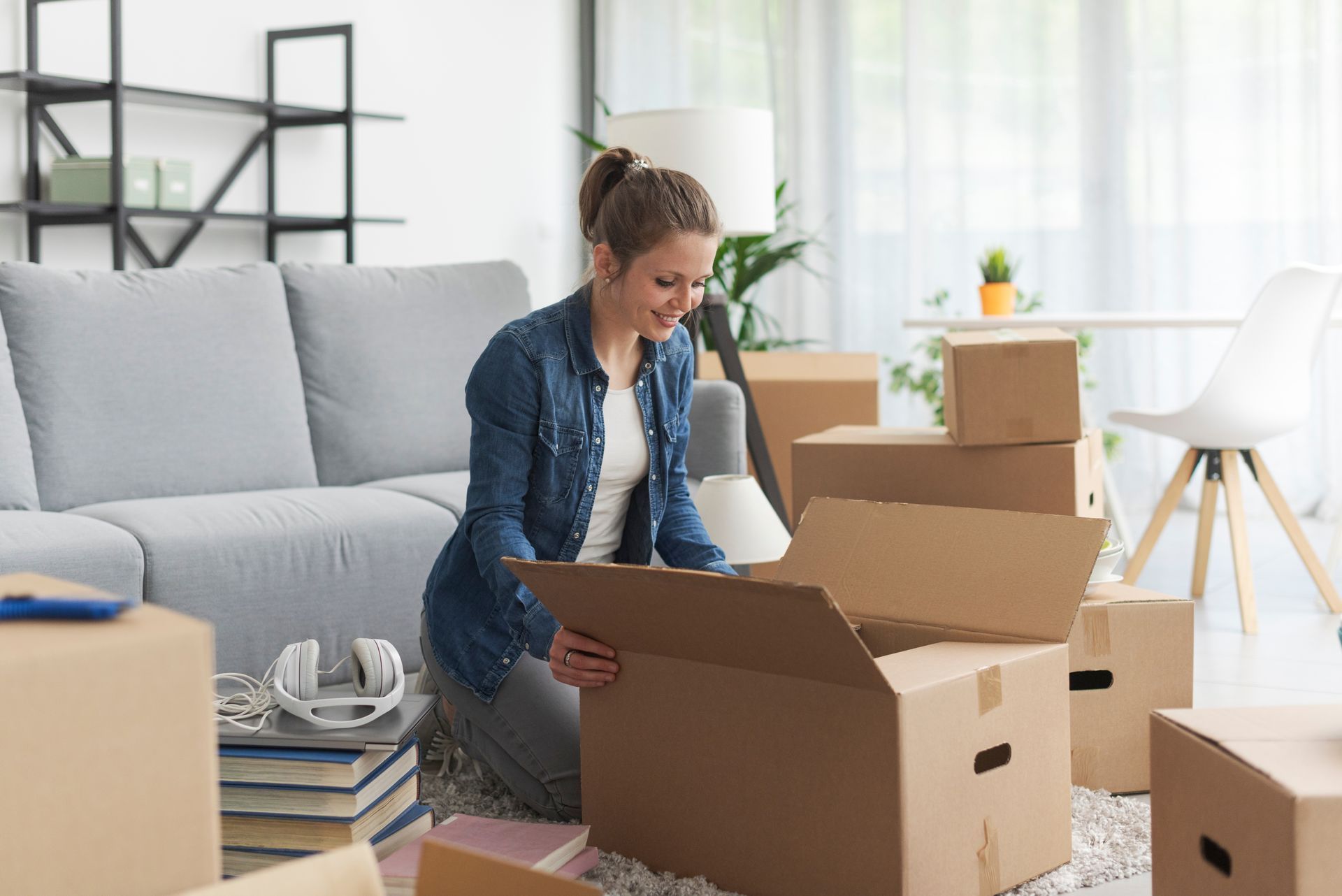 Woman unpacking boxes in a living room, smiling. Boxes and furniture surround her.