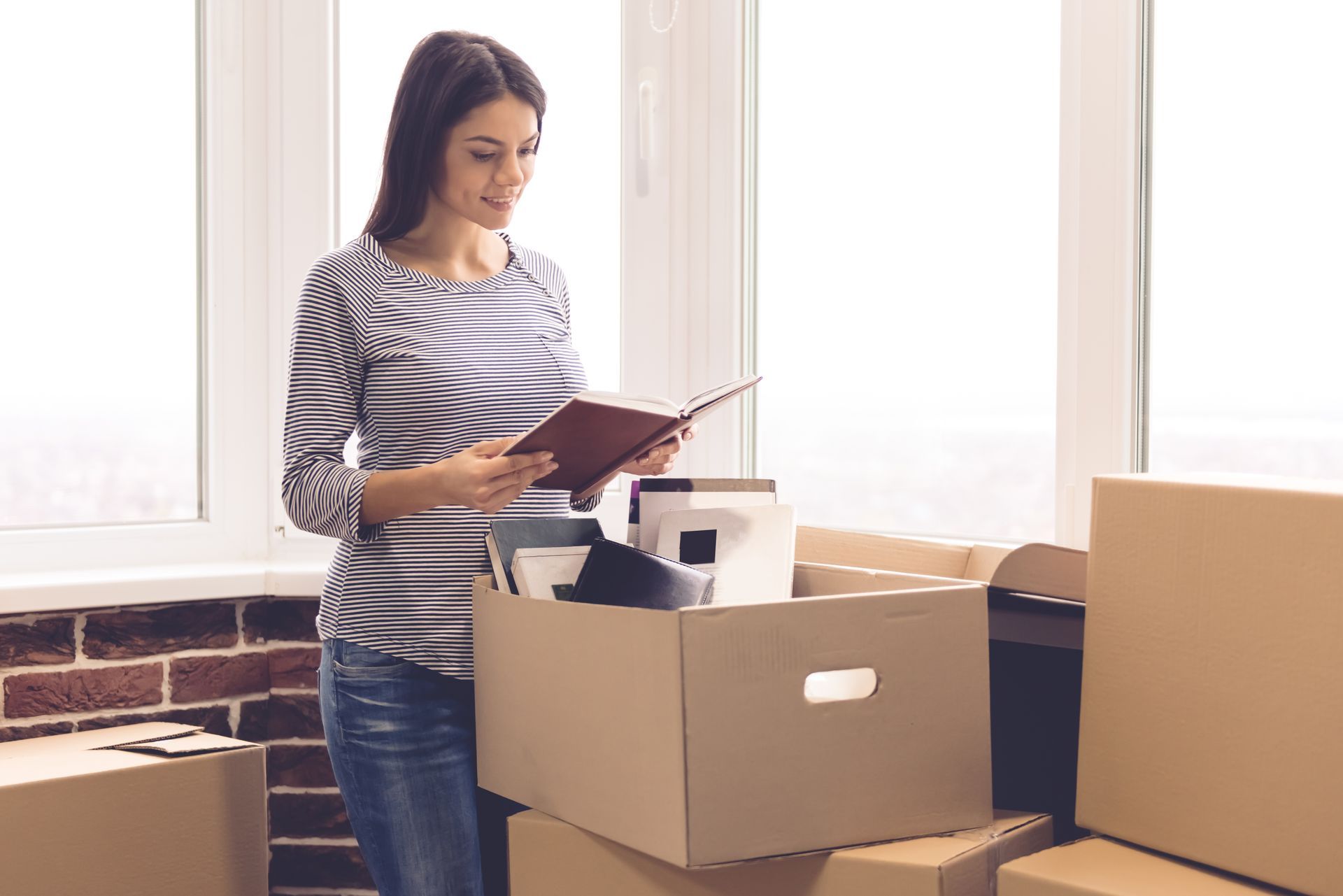 Woman reads a book near a window, surrounded by cardboard moving boxes.