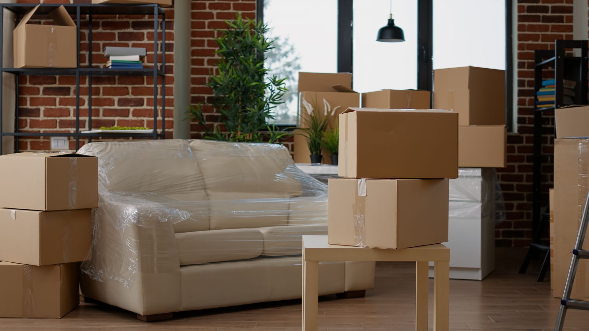 Boxes stacked in a room with a wrapped couch, shelves, and a window; moving day.