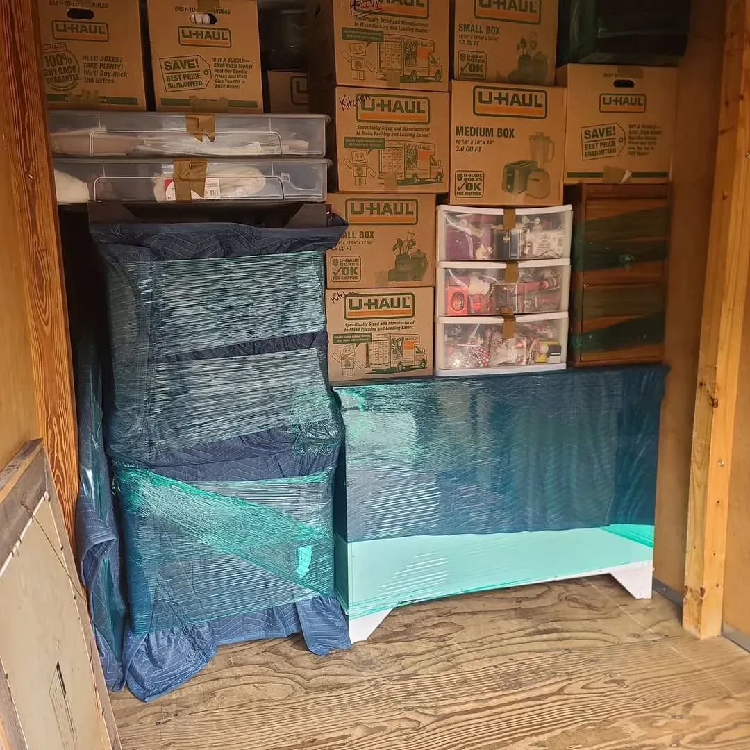 Inside a moving truck with stacked cardboard boxes and furniture wrapped in blue plastic bins.
