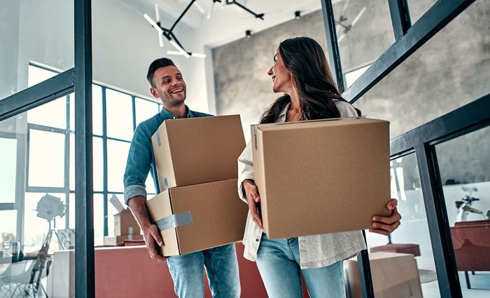 Couple carrying cardboard boxes, smiling, moving into a new home.