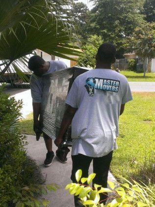 Two people carrying a large object, possibly furniture, on a sidewalk, in front of a house. One person wears a 