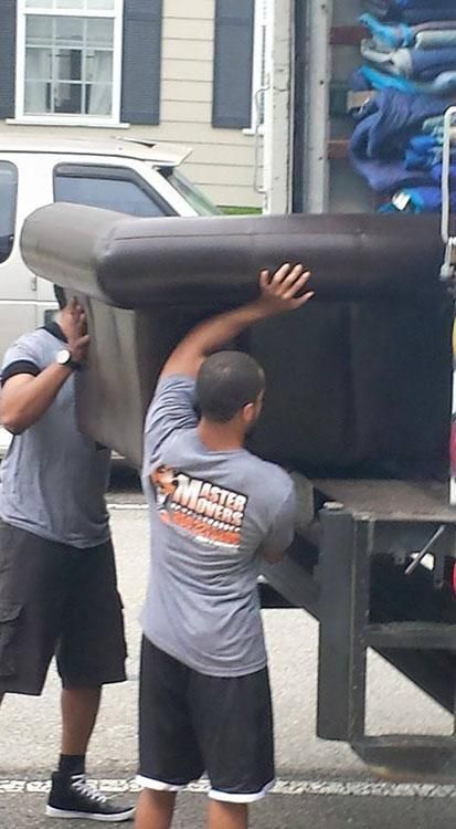 Two movers carrying a dark-colored sofa into a moving truck. The movers wear gray shirts, shorts, and sneakers.