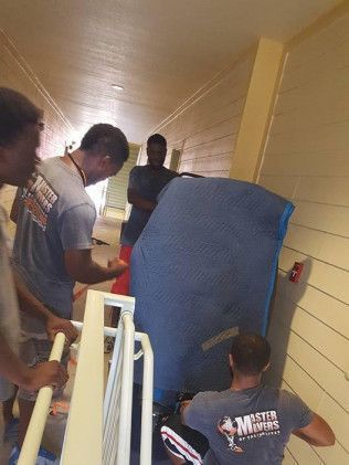 Movers carrying a large blue-wrapped object up a narrow, light-yellow hallway staircase.