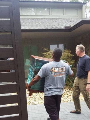 Two men carrying furniture into a house. One man is in a grey shirt, the other in a black shirt.
