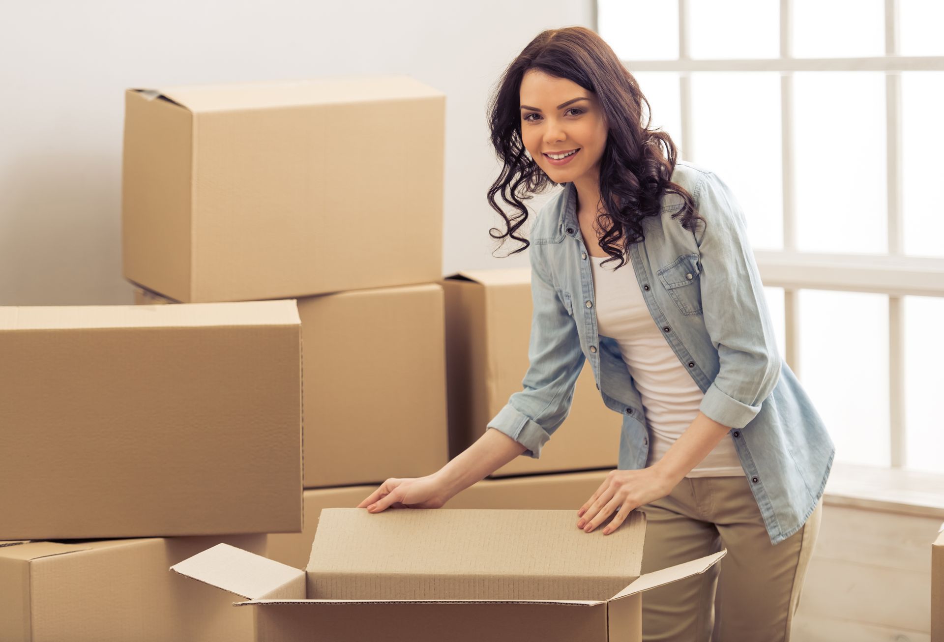 Woman packing cardboard boxes, smiling in a room with a window, wearing a denim shirt and khaki pants.
