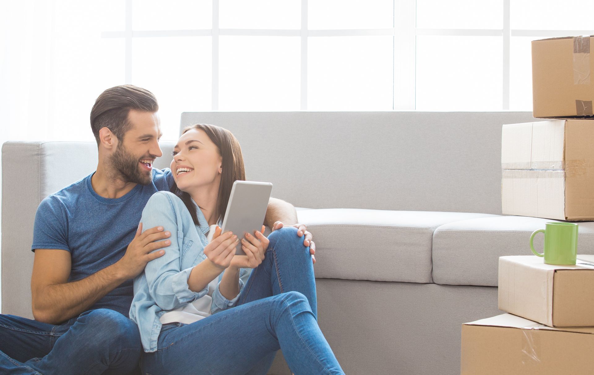Couple sitting on the floor with tablet, smiling, surrounded by moving boxes, near a couch.