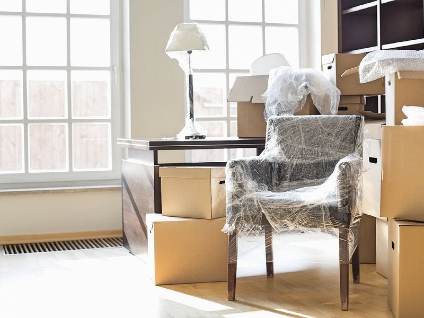 Room with packed boxes and wrapped chair; desk and lamp visible by a large window.