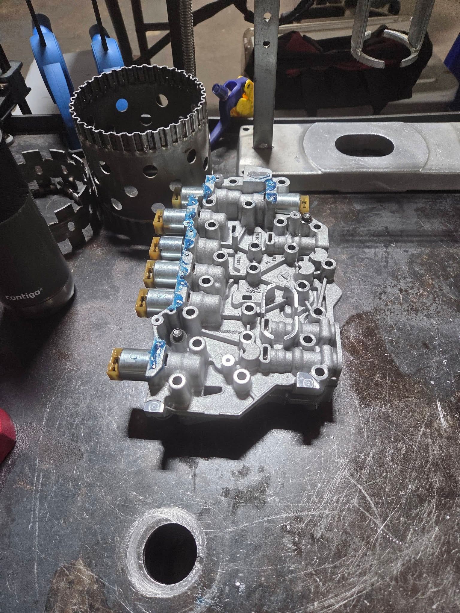 Automotive engine valve body on a workbench, with blue fittings and tools in a workshop.