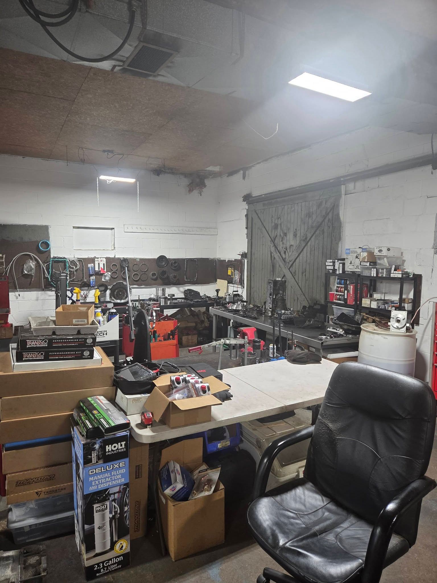 Cluttered workshop with tools, boxes, shelves, and a black office chair under bright ceiling lights