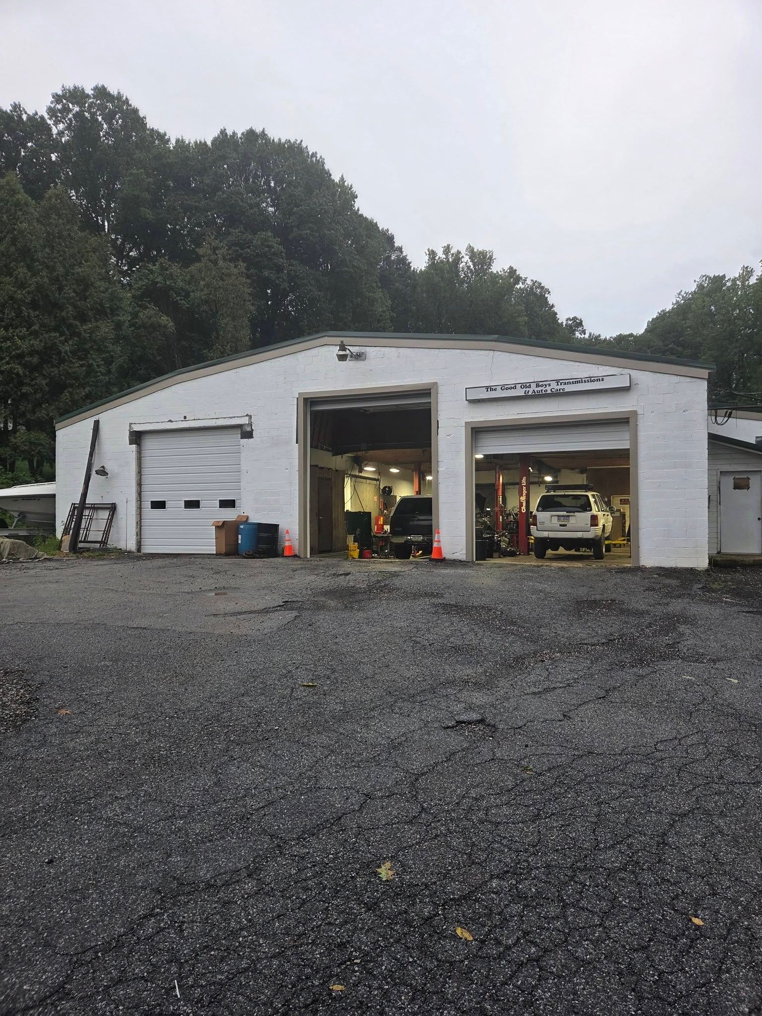 White garage/workshop with open bays, parked trucks inside, on a gravel lot surrounded by trees.