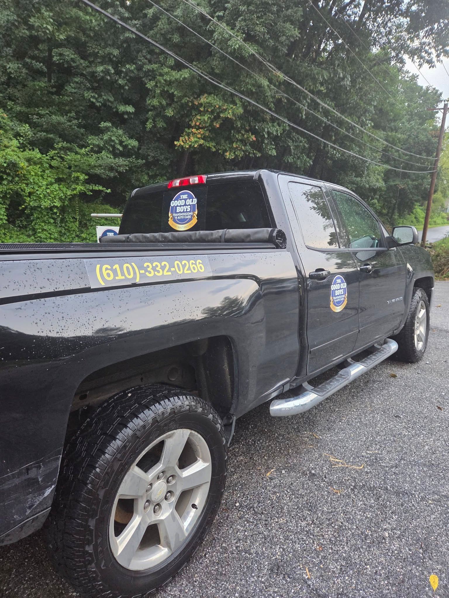 Black pickup truck parked on a gravel road beside trees, with a yellow phone number on the side.