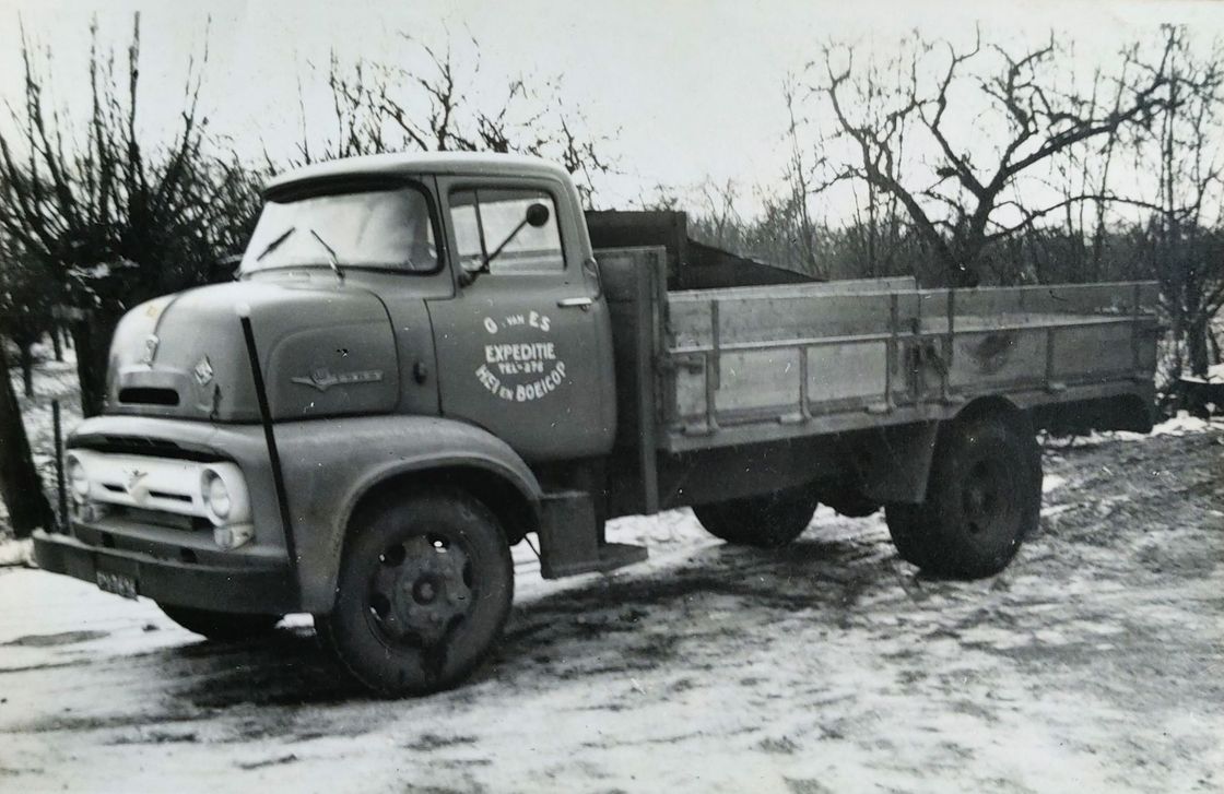 Zwart-witfoto van een oude vrachtwagen met open laadbak, geparkeerd op een besneeuwde weg in een landelijk gebied.