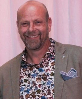 Een lachende, kale man in een bloemenhemd en een grijze blazer met pochet staat binnen.
