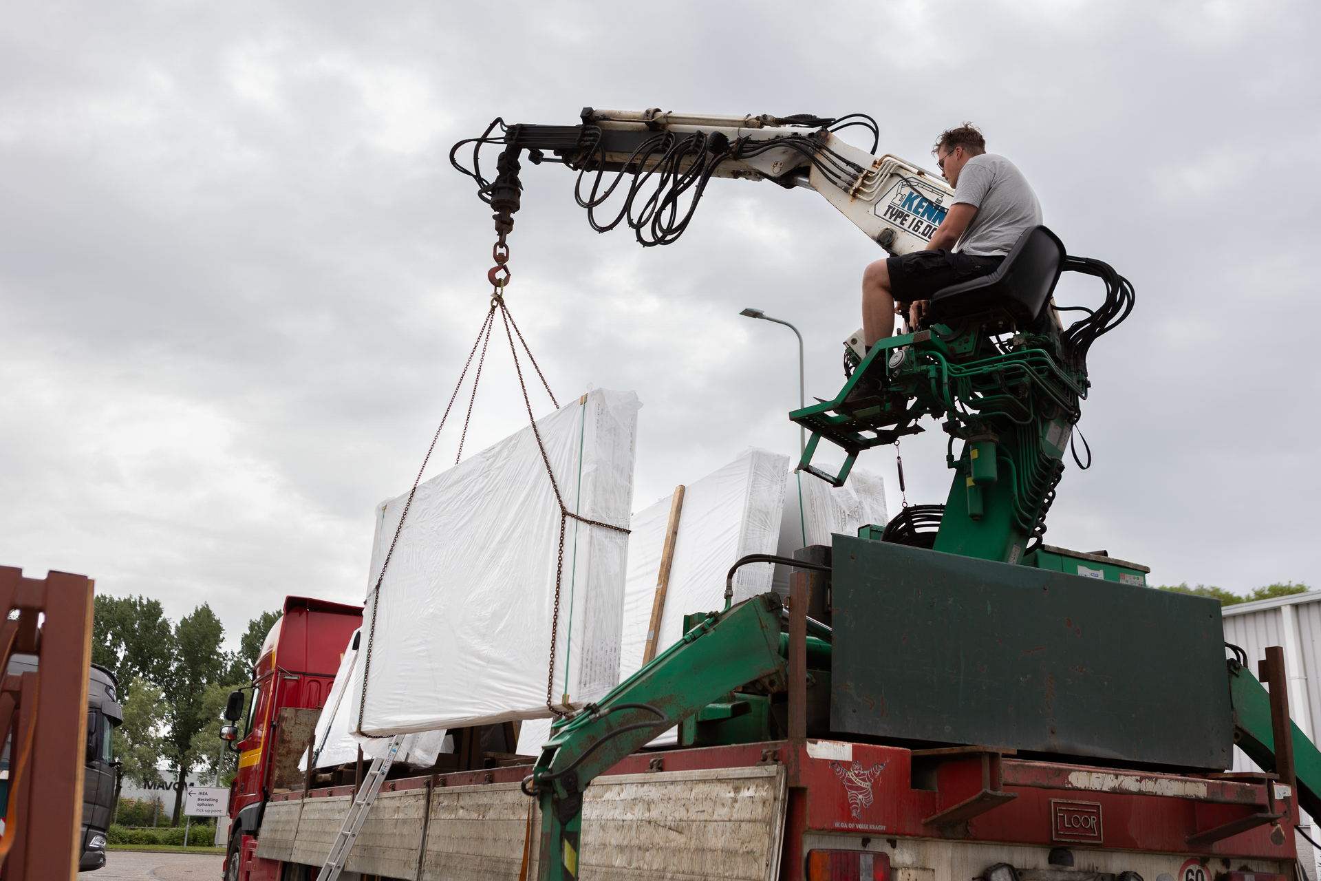 Een medewerker bedient een groene kraan waarmee hij een wit paneel op de laadbak van een vrachtwagen tilt.