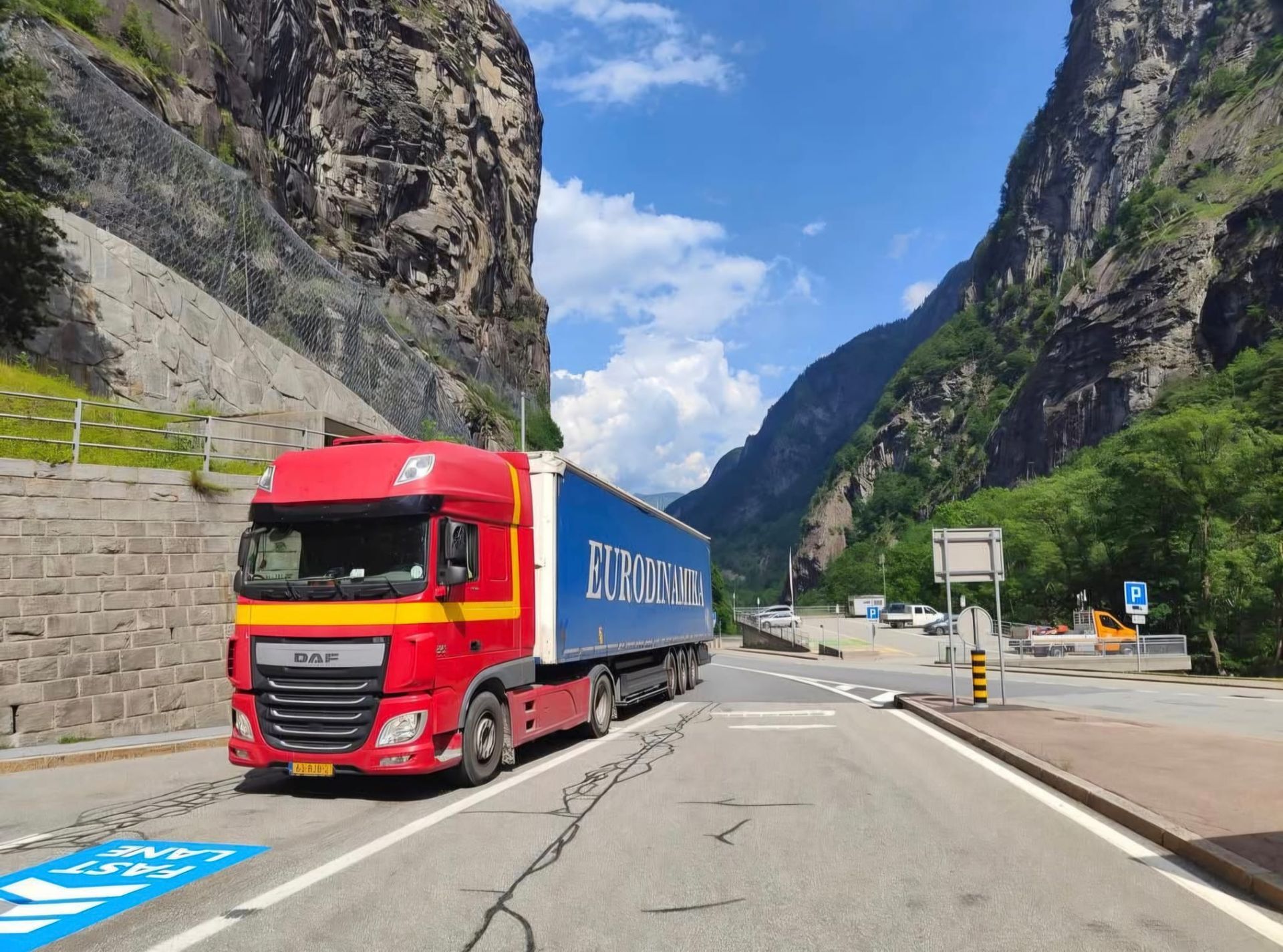 Een rode vrachtwagen rijdt door een bergkloof over een kronkelende weg onder een blauwe hemel.