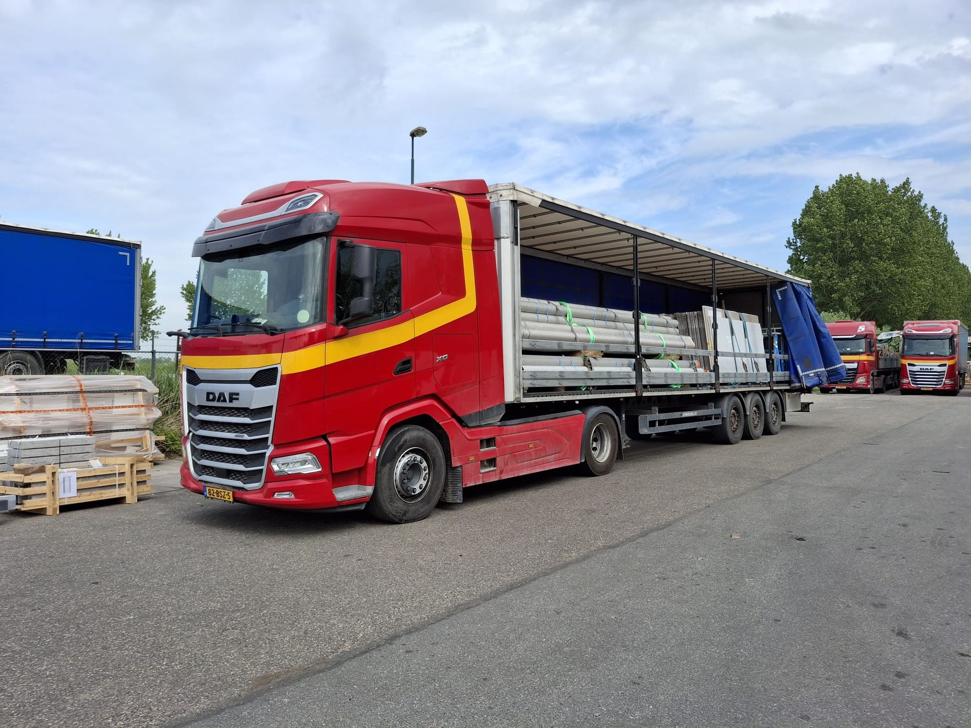 Rode vrachtwagen met open oplegger, geladen met ingepakte goederen, geparkeerd op een industrieterrein.