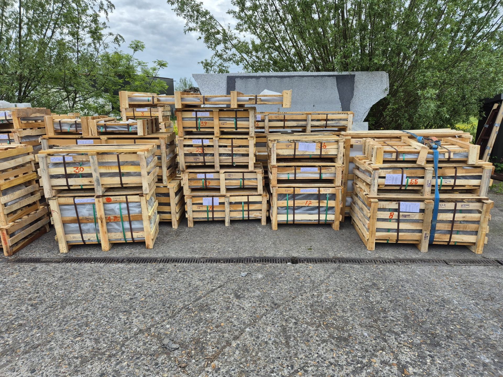Stapels houten pallets en kratten op een grindterrein, met bomen en een gebouw op de achtergrond.