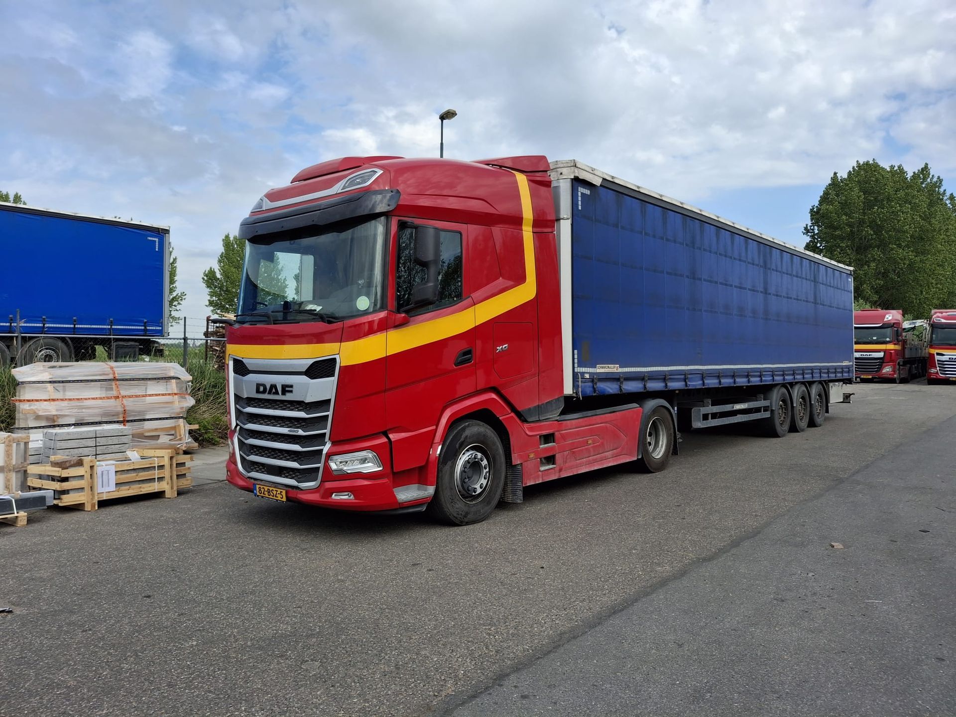 Een rode vrachtwagen met blauwe oplegger staat geparkeerd op een terrein naast gestapelde pallets onder een bewolkte hemel.