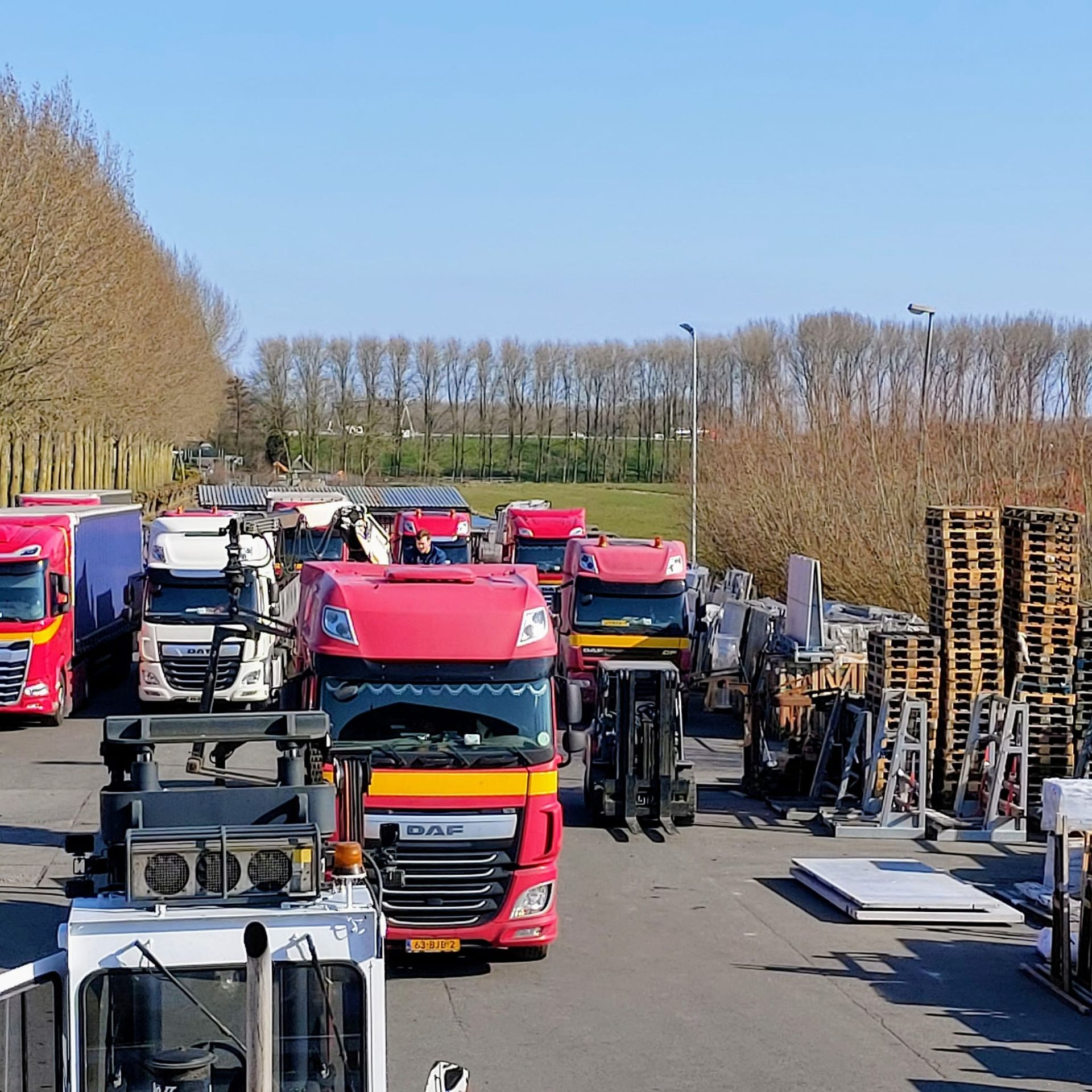 Een konvooi vrachtwagens staat opgesteld langs een weg naast opgestapelde pallets onder een heldere blauwe hemel.