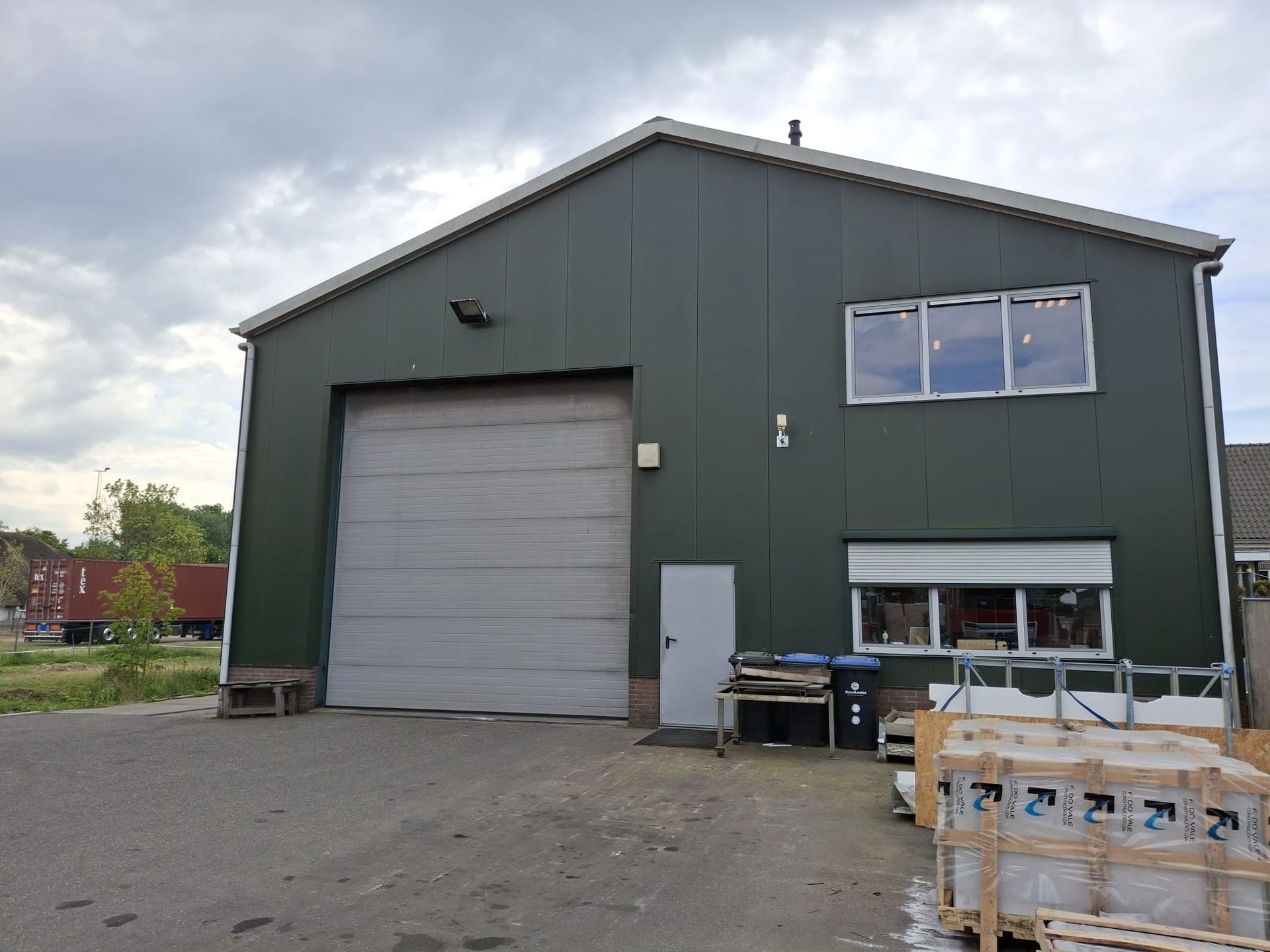 Groen industrieel gebouw met een grote garagedeur, ramen en bouwmaterialen aan de buitenkant.