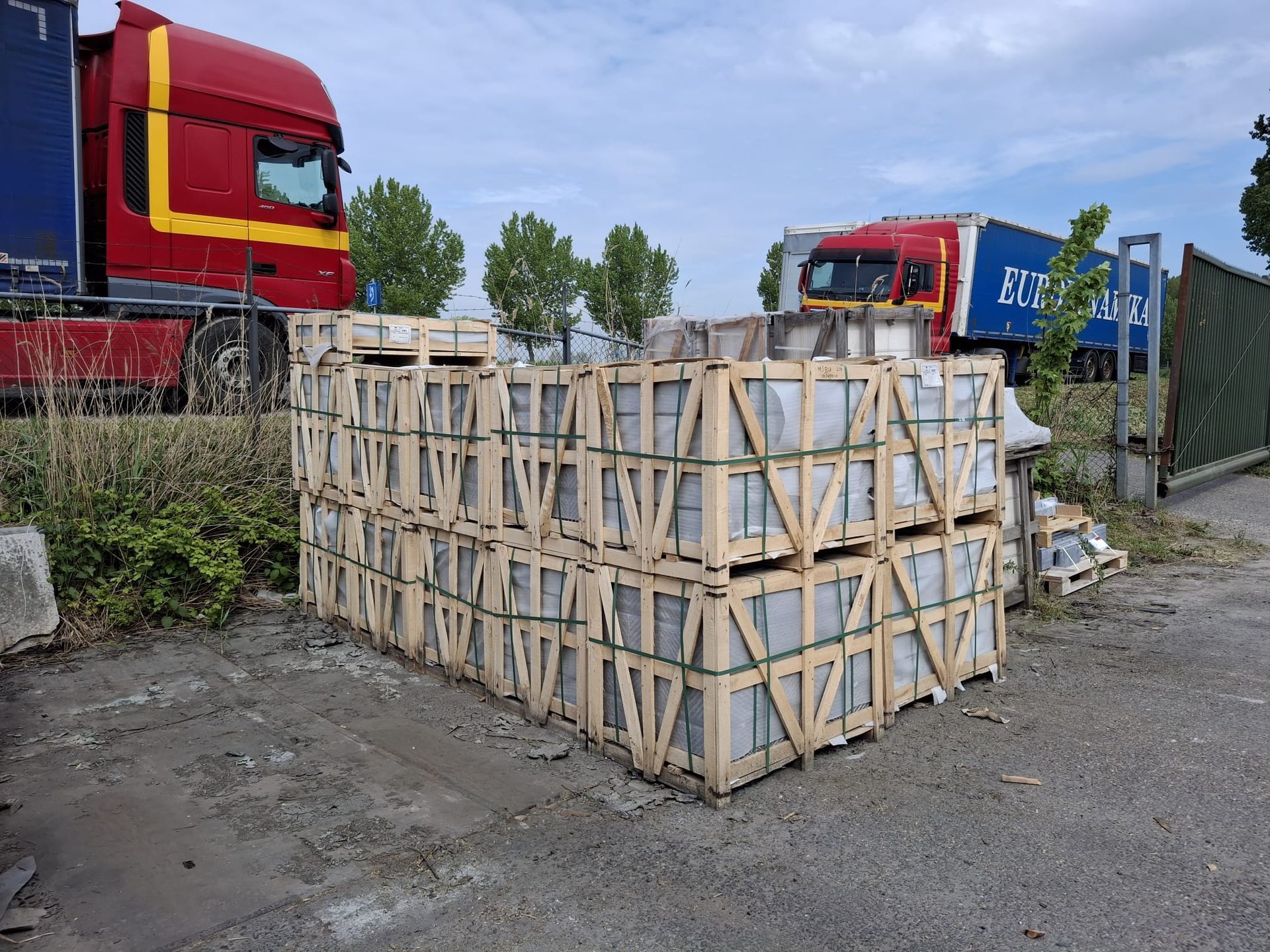 Houten kratten vastgebonden op een pallet, vlakbij rode vrachtwagens op een industrieterrein.