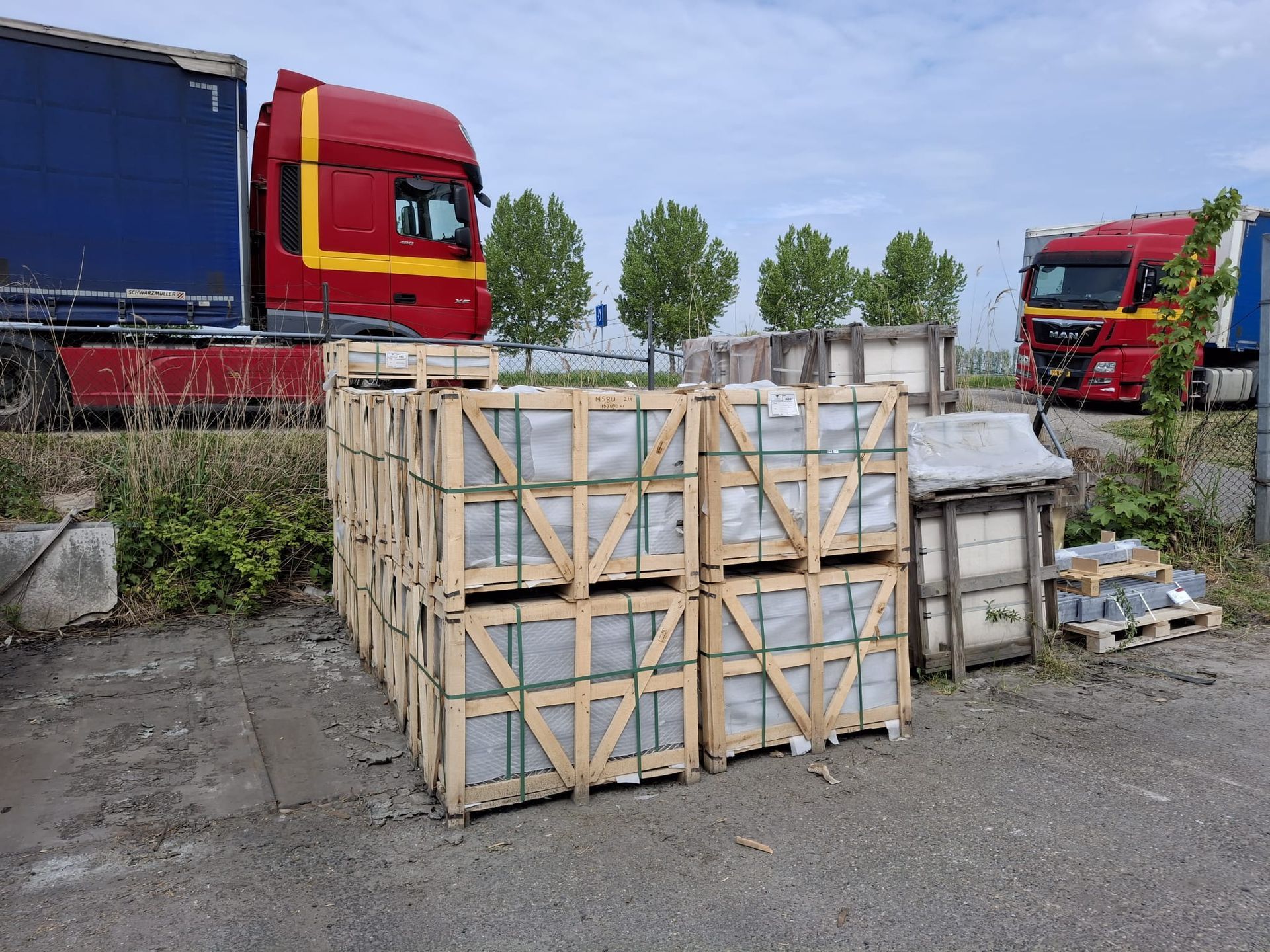 Pallets met houten kratten naast rode vrachtwagens op een industrieterrein.