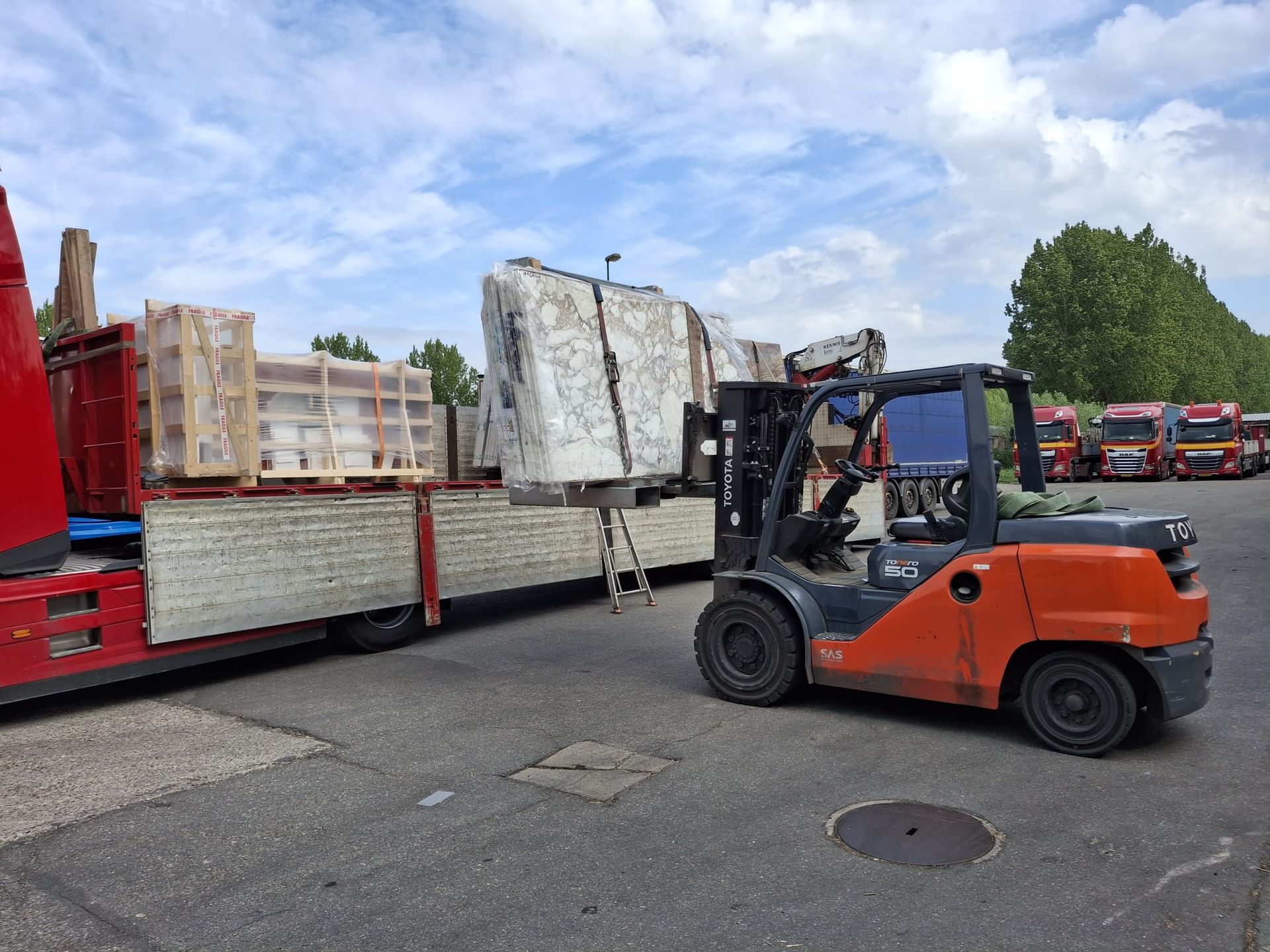 Een oranje heftruck tilt stenen platen op naast een rode vrachtwagen op een buitenterrein.