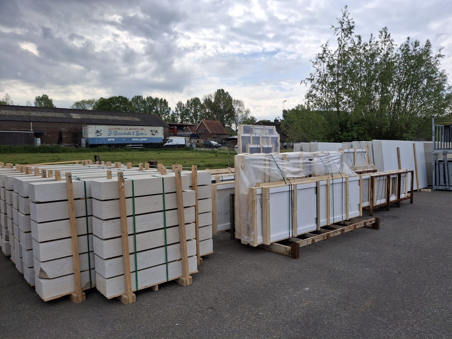 Stapels ingepakte witte pallets op houten sleden op een industrieterrein in de openlucht, onder een bewolkte hemel.
