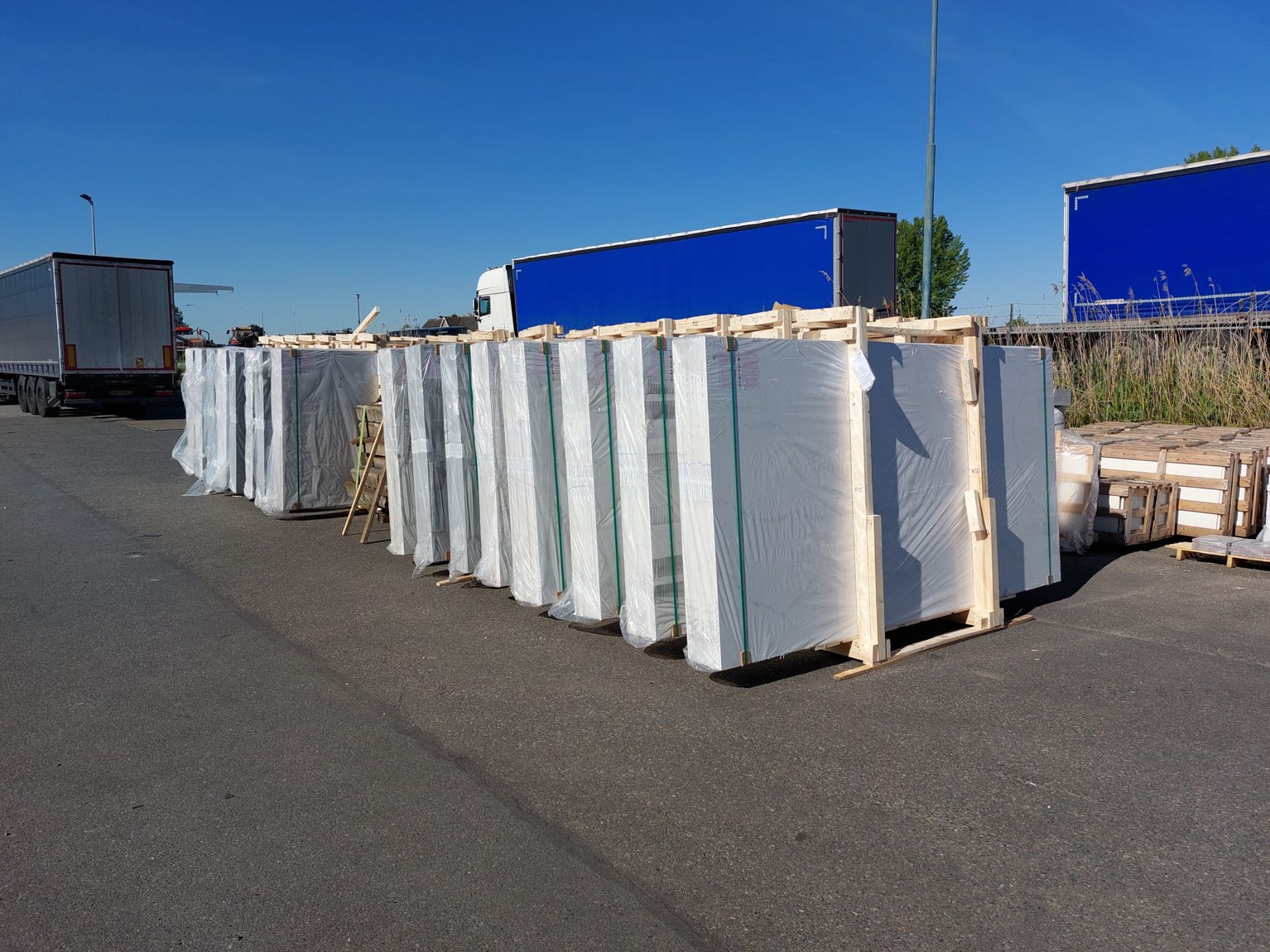 Witte zeecontainers staan op een geplaveid terrein naast blauwe trailers onder een heldere hemel.