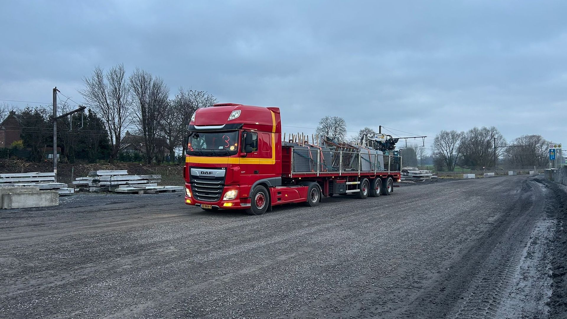 Rode vrachtwagen met oplegger geparkeerd op een grindterrein onder een bewolkte hemel.