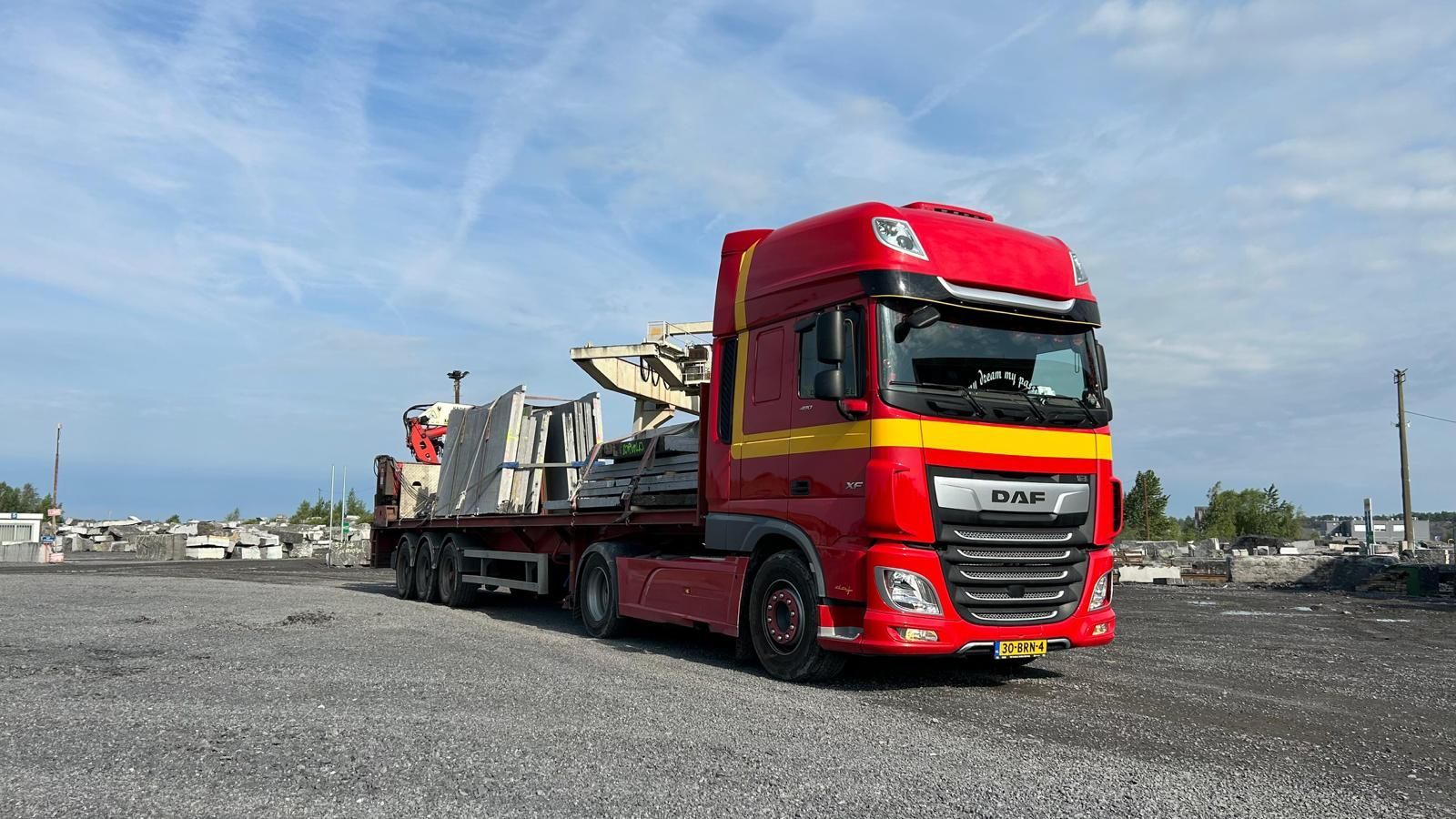 Een rode vrachtwagen met oplegger, waarop machines staan ​​geparkeerd op een grindpad onder een blauwe hemel.